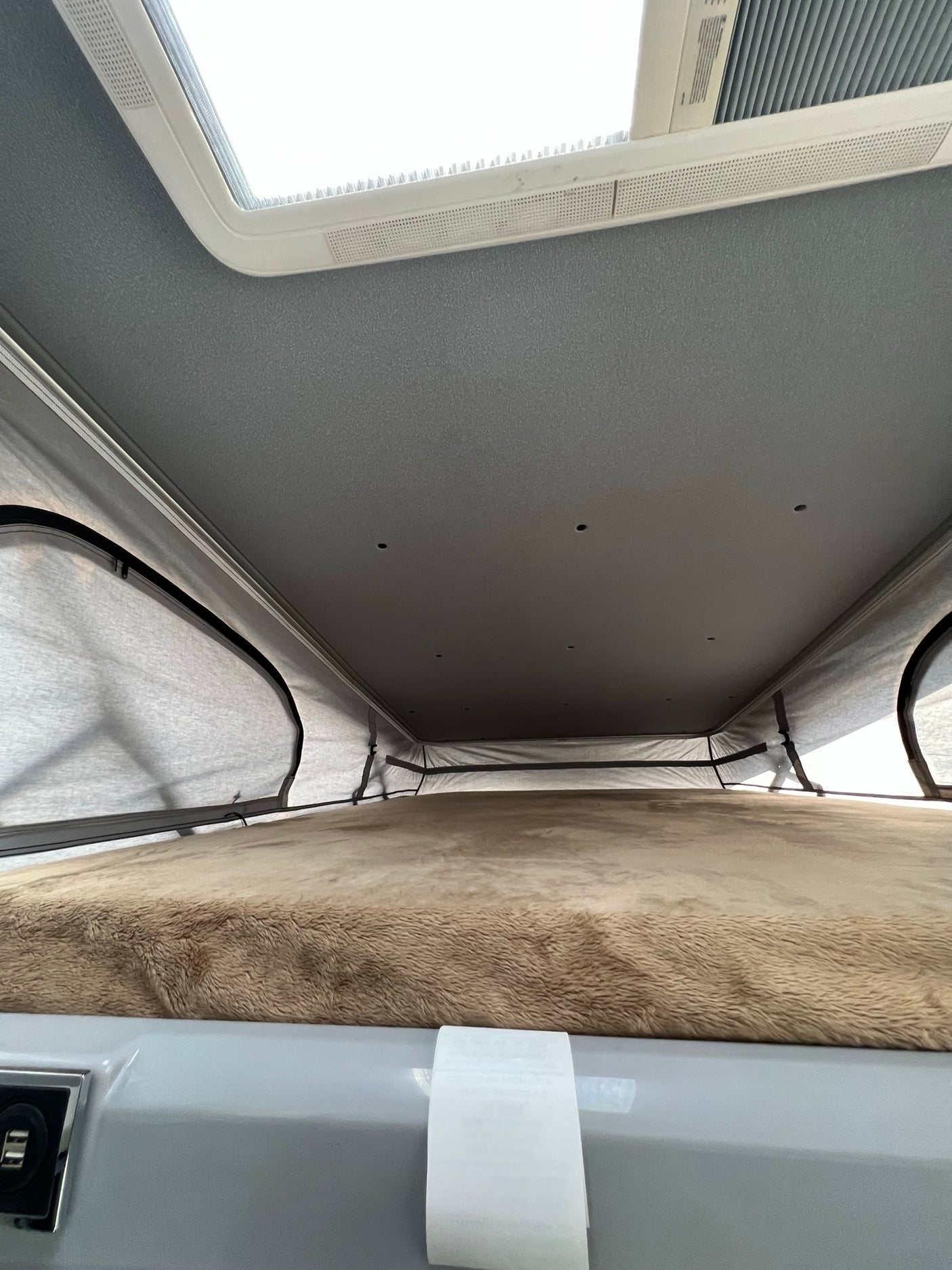 RV rental interior: Upward view of a pop-top sleeping area with furry bed, soft tent walls, ceiling lights, and skylight.