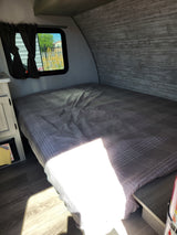 Cozy RV rental interior: bedroom with plaid bed, window, and wood-look walls. Ideal for travel and comfortable stays.