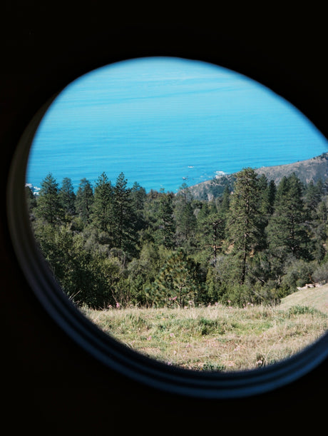 Stunning coastal view from an RV rental's round window: blue ocean, green mountains, and wildflowers on a grassy hill.