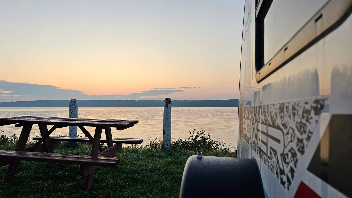 Golden hour at a lakeside RV rental spot. An RV on the right, picnic tables on the left, facing a calm lake and colorful sky.