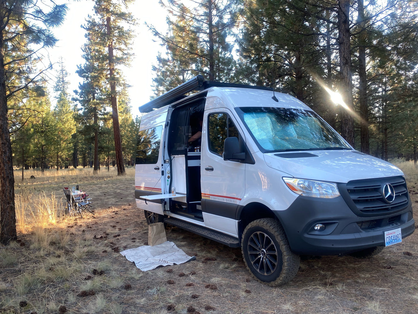 A Storyteller Overland Mercedes Sprinter rv rental camper van is parked in a sunny forest, side door open for camping.