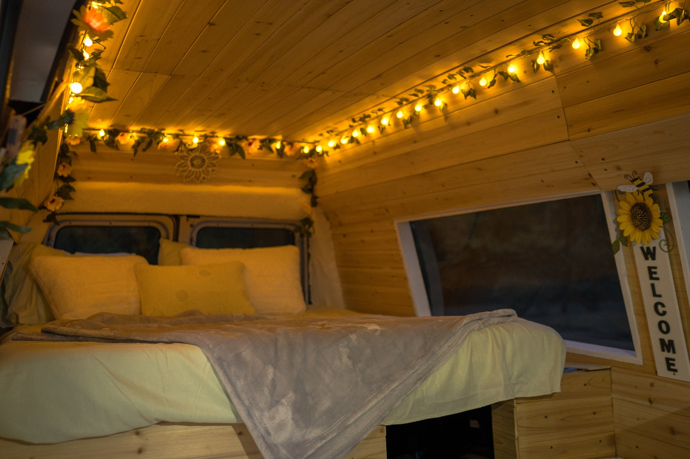 Cozy RV rental bedroom with warm wood paneling, charming fairy lights, and a "WELCOME" sign. Ideal for glamping.