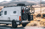 This white Sprinter RV rental is prepped for adventure, featuring a bike rack, spare tire, & roof rack in scenic terrain.