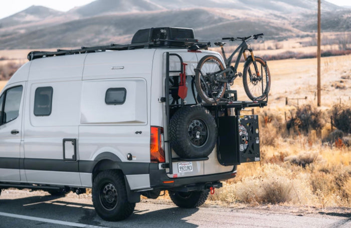 This white Sprinter RV rental is prepped for adventure, featuring a bike rack, spare tire, & roof rack in scenic terrain.