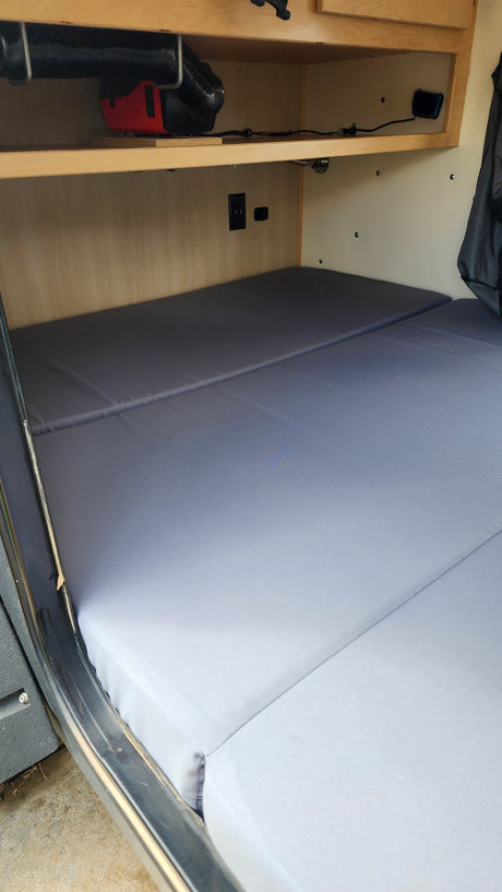 Inside an RV rental: A simple bed with gray mattresses, wooden shelves, and a heater/ventilation system.