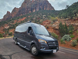 Dark grey Sprinter RV rental parked on a scenic road with majestic red rock mountains and green vegetation.