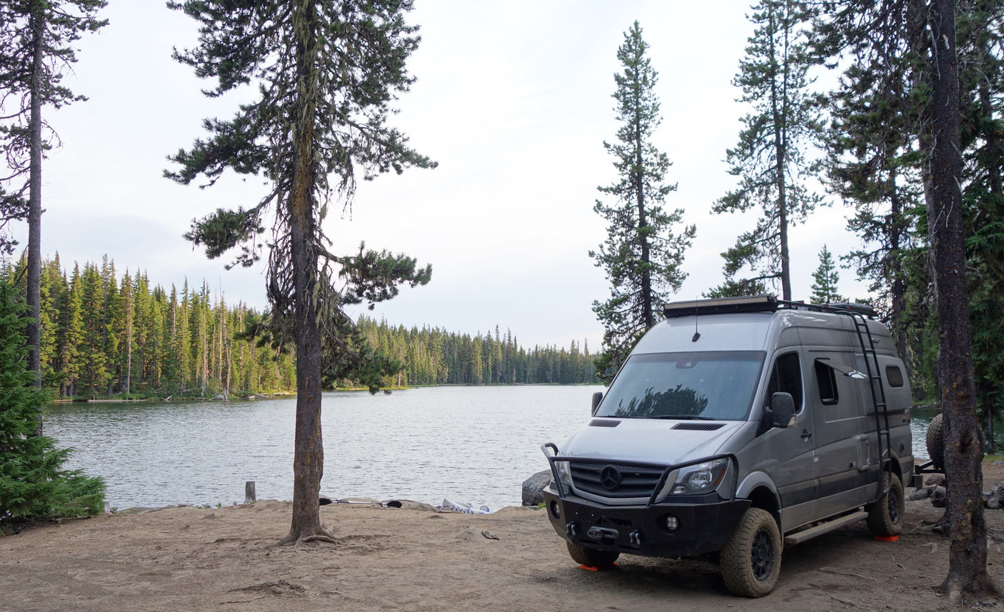 Lakeside camping with an RV rental. A gray custom camper van is parked on dirt next to a calm lake and dense forest.