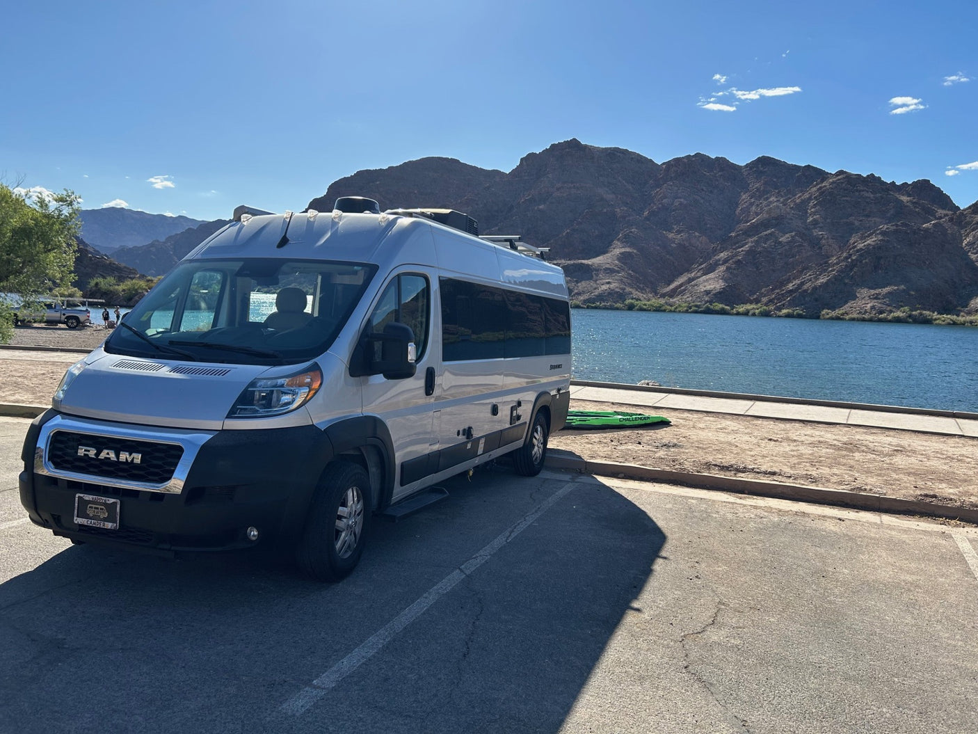 Silver rv rental van parked next to a blue lake and rugged mountains, under a bright sky. Perfect for outdoor travel.