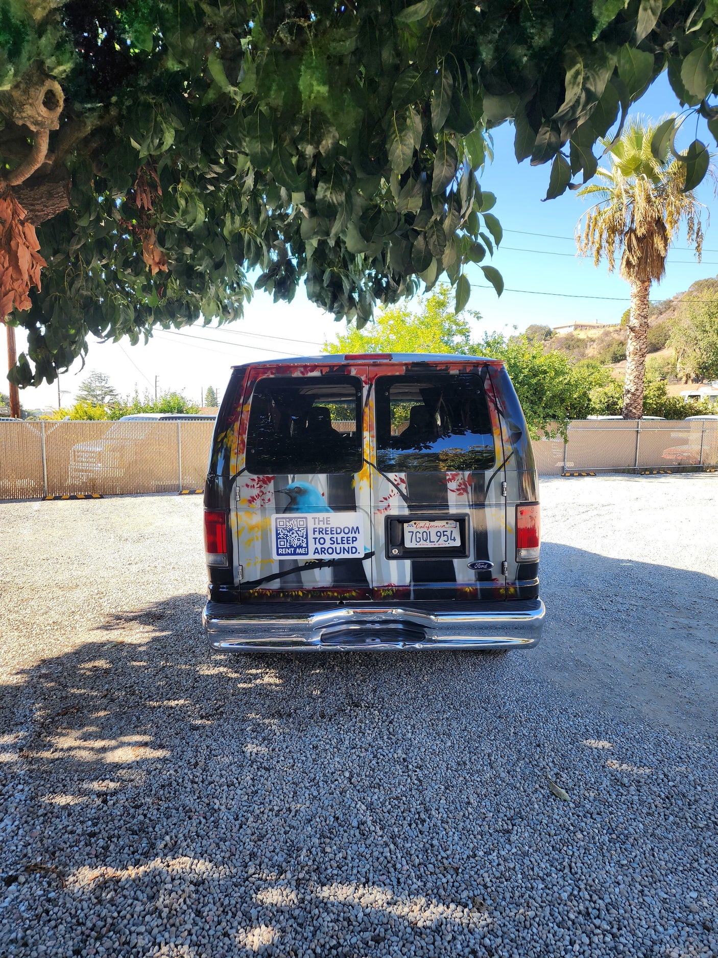 Rear view of a custom RV rental van with a "RENT ME!" QR code, parked on a gravel lot under a tree.