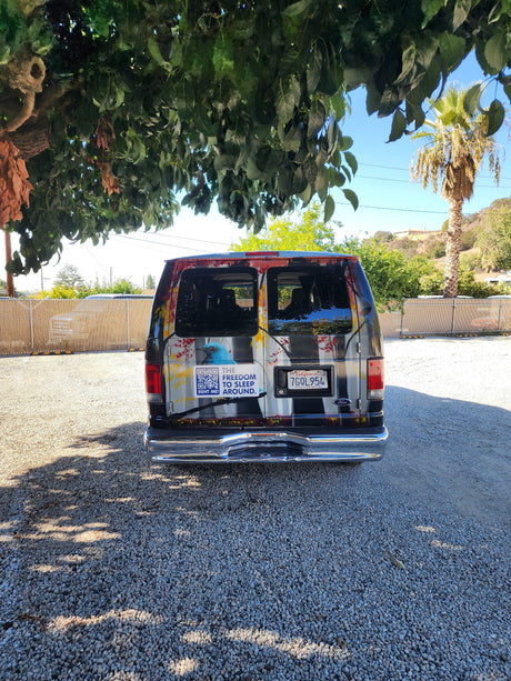 Rear view of a custom RV rental van with a "RENT ME!" QR code, parked on a gravel lot under a tree.