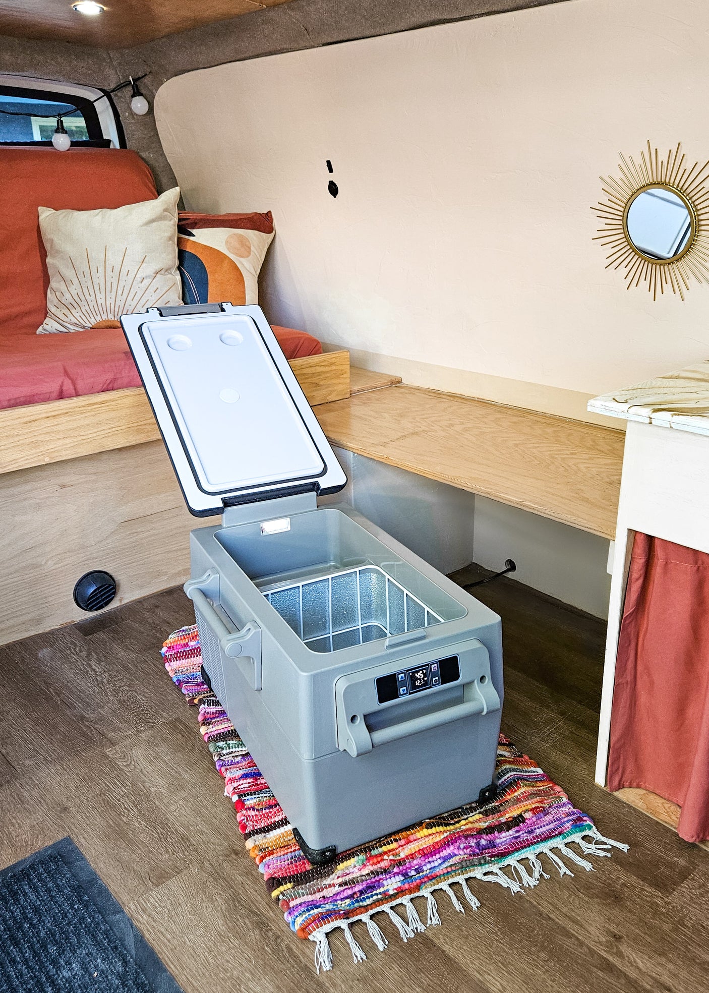 Spacious RV rental interior featuring a portable fridge on a colorful rug, wooden bed, and shelving for a comfy road trip setup.