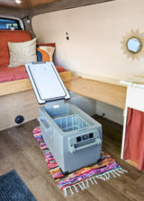 Spacious RV rental interior featuring a portable fridge on a colorful rug, wooden bed, and shelving for a comfy road trip setup.