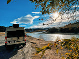 Explore with an RV rental! White van by a sunlit river, autumn leaves, and distant snowy hills under blue sky.