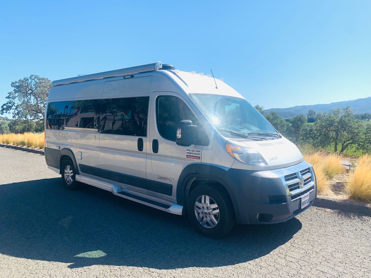 This silver van-style RV rental is parked on a paved road, bathed in sunlight against a backdrop of green hills and golden grass.