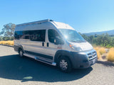 This silver van-style RV rental is parked on a paved road, bathed in sunlight against a backdrop of green hills and golden grass.