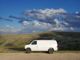 White camper van, resembling an RV rental, parked on a dirt road with expansive cloudy mountain views.