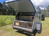 This rv rental, a grey teardrop camper, showcases its open rear hatch revealing a fully equipped outdoor kitchen setup.