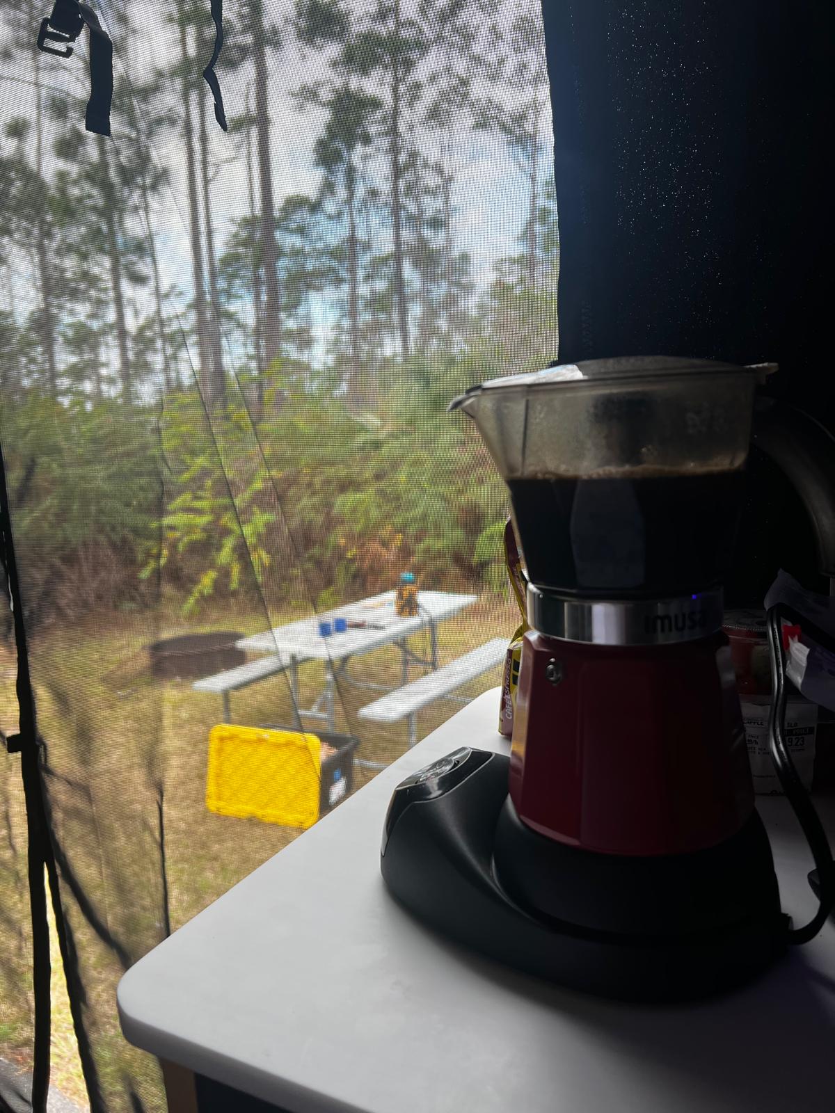 Fresh coffee brewing in an electric Moka pot inside an **rv rental**, overlooking a peaceful campsite with pine trees & picnic table.