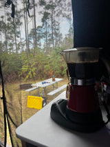 Fresh coffee brewing in an electric Moka pot inside an **rv rental**, overlooking a peaceful campsite with pine trees & picnic table.