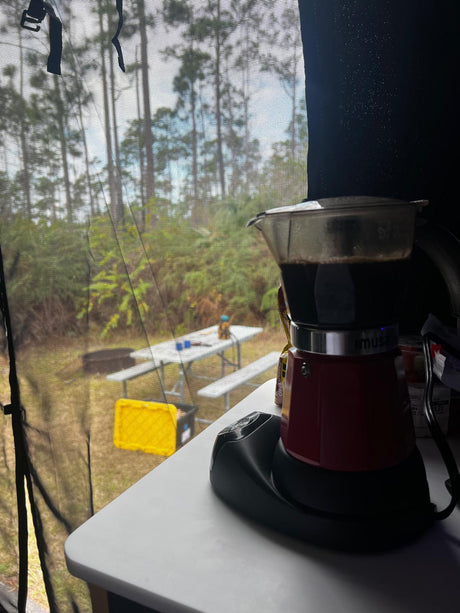 Fresh coffee brewing in an electric Moka pot inside an **rv rental**, overlooking a peaceful campsite with pine trees & picnic table.