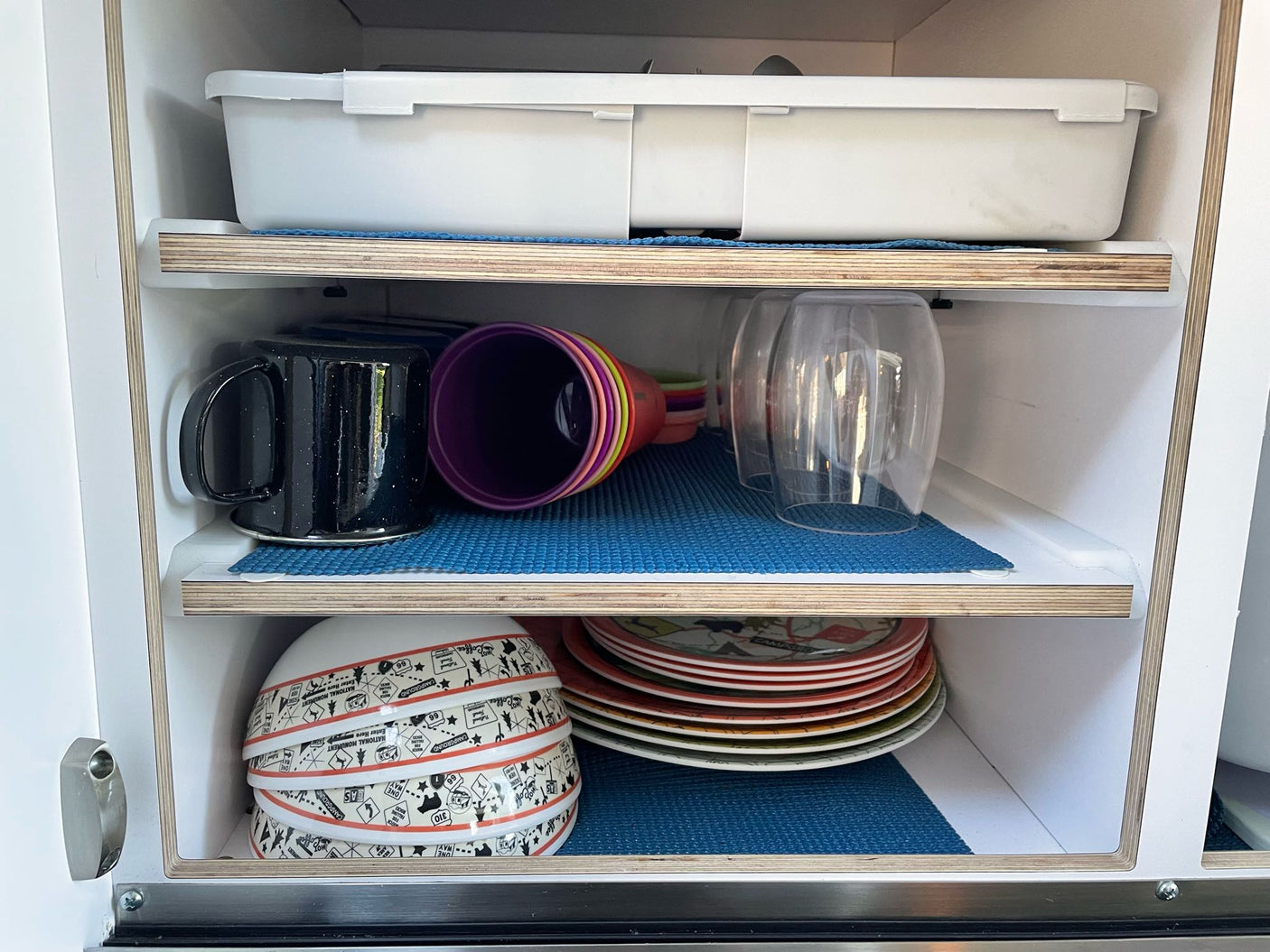 Organized RV rental kitchen cabinet with dishes, mugs, cups, and bowls on blue mats, perfect for travel.