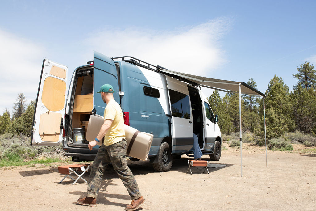 Man unpacks an RV rental van, a blue and white camper with an awning extended, for an outdoor adventure.