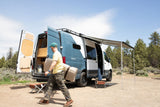 Man unpacks an RV rental van, a blue and white camper with an awning extended, for an outdoor adventure.