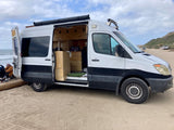 Converted camper van, resembling an RV rental, parked on sandy beach. Open door reveals a custom interior, ready for travel adventures by the ocean.