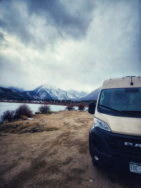A beige RV rental van is parked on a dirt shore next to a lake, with snow-capped mountains under a cloudy sky.