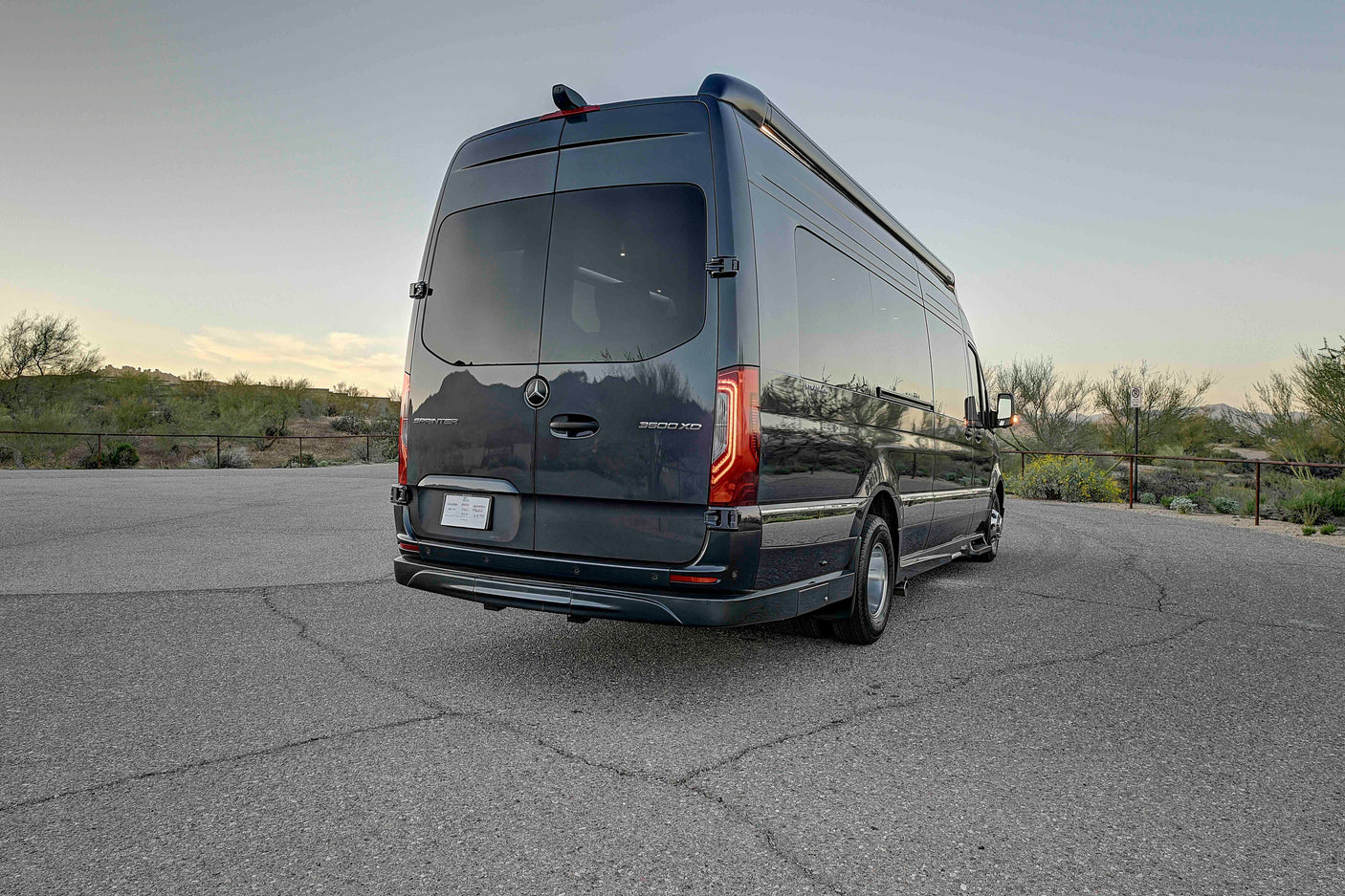 Dark grey Mercedes Sprinter RV rental, seen from the rear-side, parked on asphalt with a desert landscape. Ideal for travel.