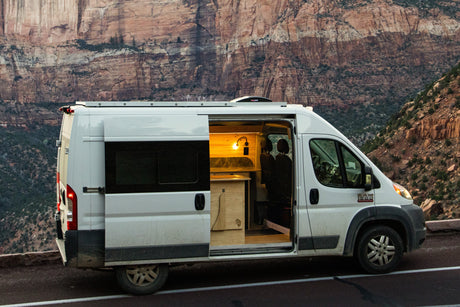 A white rv rental camper van, with its door open revealing a warm interior, parked on a road beside towering red cliffs.