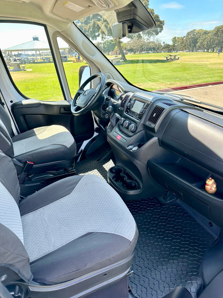 Inside this RV rental: front cabin, seats, dashboard, and a clear view of a park with a gazebo and trees.