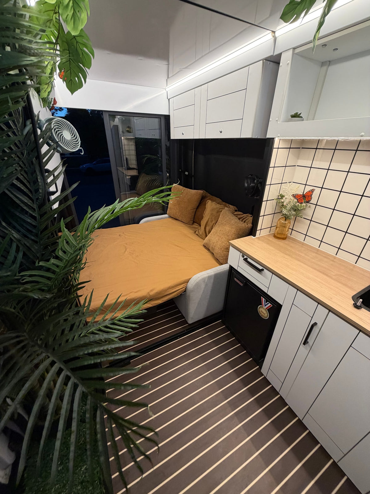 RV rental interior featuring a neatly made bed, compact kitchen with wood counter, and green plant decor. Modern, cozy living. — photo 6