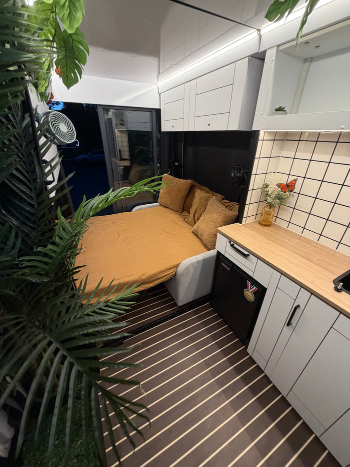 RV rental interior featuring a neatly made bed, compact kitchen with wood counter, and green plant decor. Modern, cozy living.