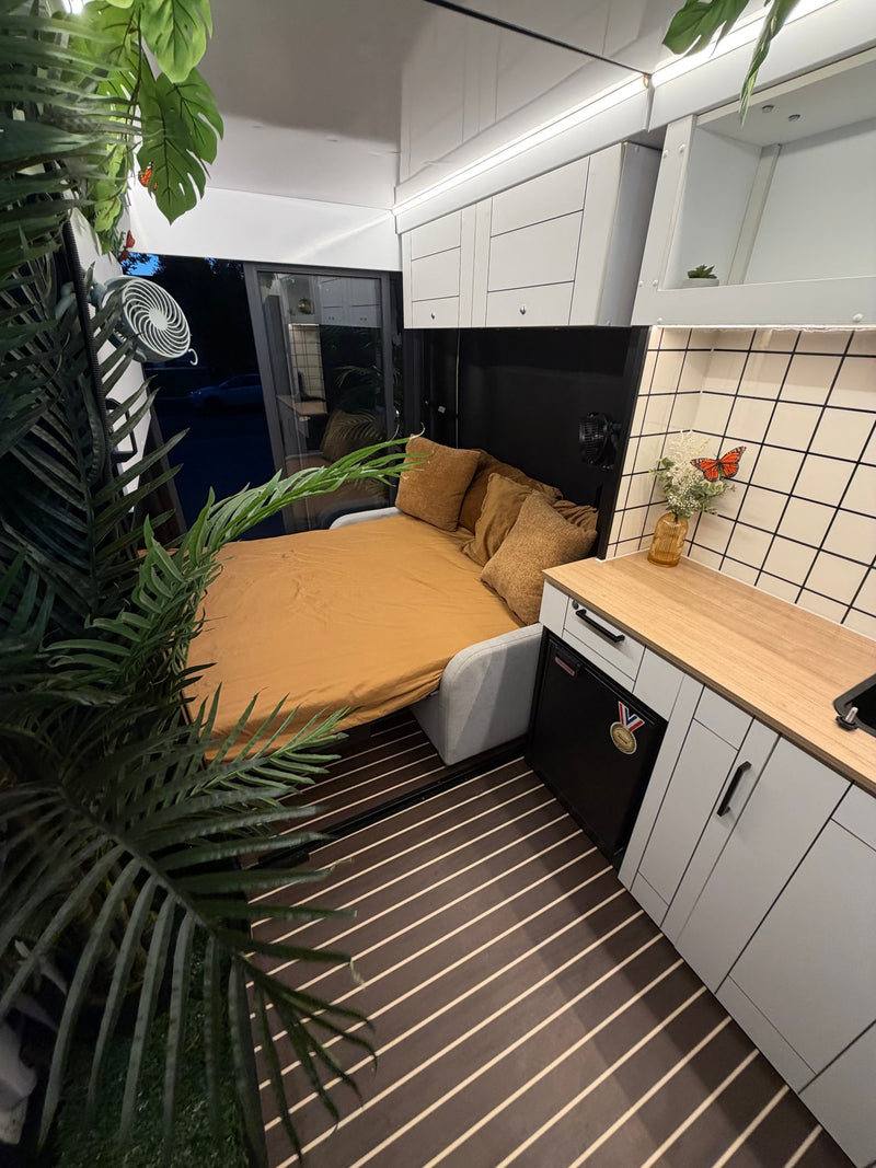 RV rental interior featuring a neatly made bed, compact kitchen with wood counter, and green plant decor. Modern, cozy living. — photo 6