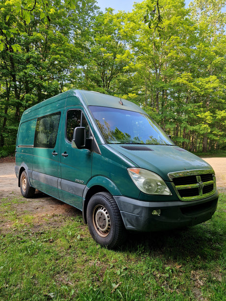 Green Dodge Sprinter camper van, great for an RV rental, parked in a sunny, tree-filled forest.