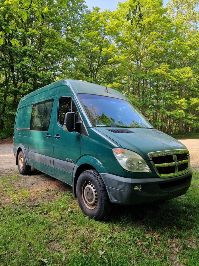 Green Dodge Sprinter camper van, great for an RV rental, parked in a sunny, tree-filled forest.