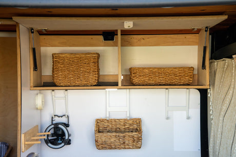 Efficient RV rental storage: overhead cabinets with wicker baskets, a fan, and organizational hooks in a cozy camper interior.