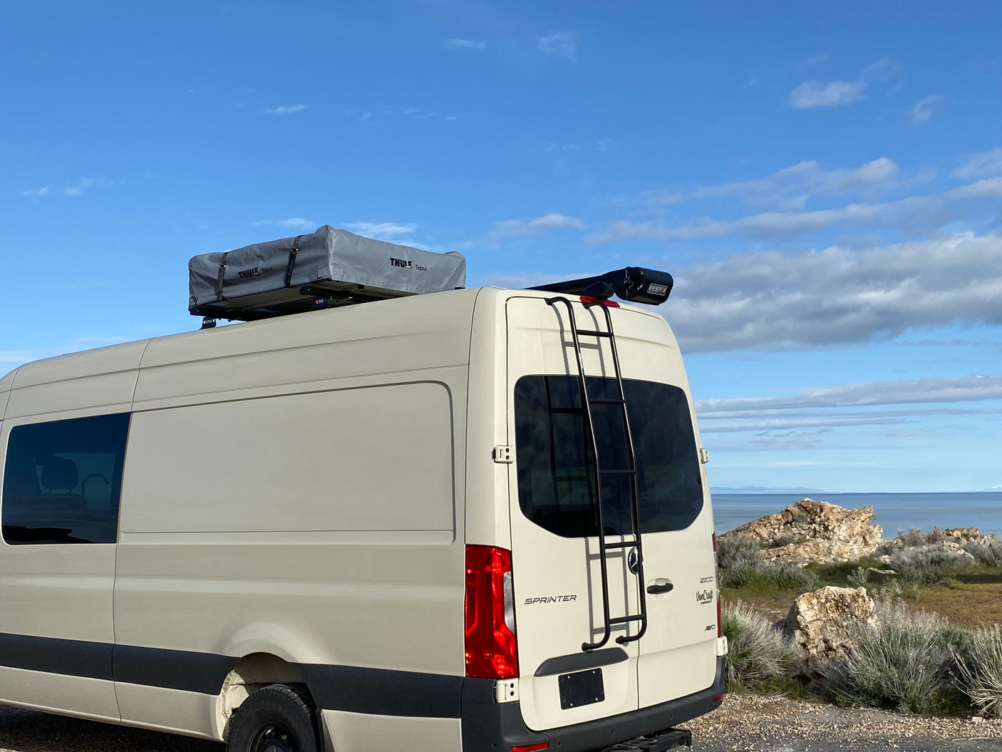 Beige Sprinter RV rental with a rooftop tent & fishing rod carrier, parked by a scenic lake/ocean for your adventure.