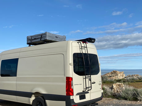 Beige Sprinter RV rental with a rooftop tent & fishing rod carrier, parked by a scenic lake/ocean for your adventure.