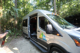 A silver WanderVans RV rental is parked in lush woods. Its open side door reveals a spacious interior for adventure.