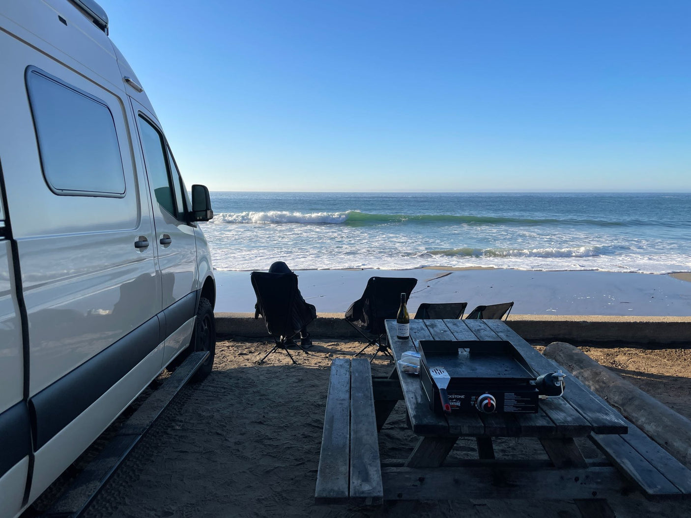 Perfect rv rental spot: white van by the beach, people enjoying waves, and a picnic table with a griddle.