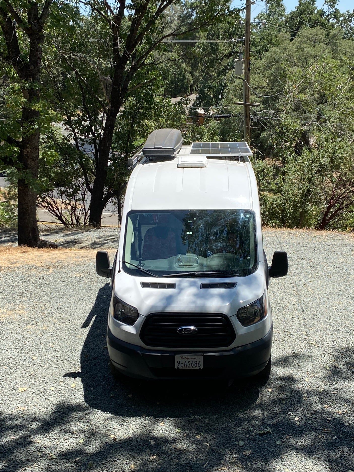 White camper van, possibly an RV rental, on a gravel lot under trees, with solar panels & rooftop cargo box.