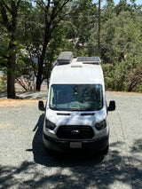 White camper van, possibly an RV rental, on a gravel lot under trees, with solar panels & rooftop cargo box.