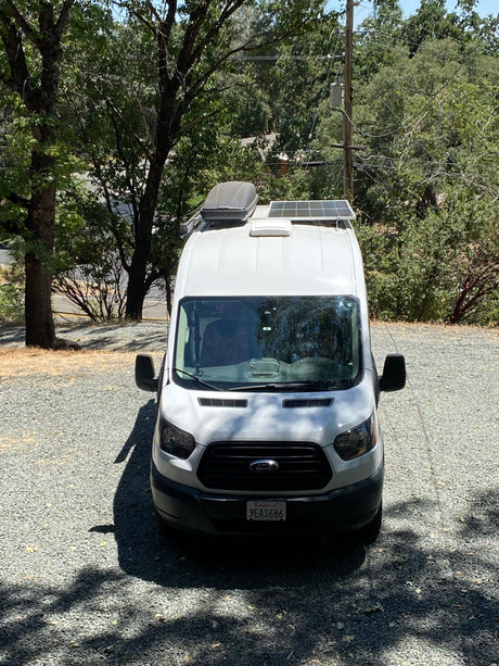 White camper van, possibly an RV rental, on a gravel lot under trees, with solar panels & rooftop cargo box.