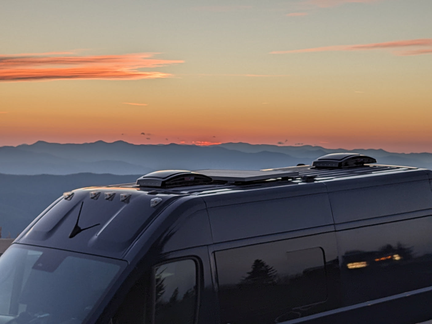 A dark RV rental, equipped with rooftop gear, overlooks a vibrant orange sunset and layered mountains at dusk.