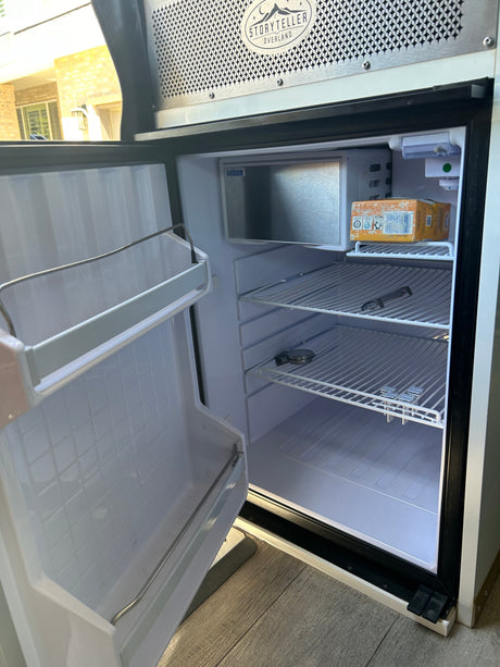 RV rental kitchen: Interior of a clean, compact refrigerator with open door, empty shelves, and a small freezer.