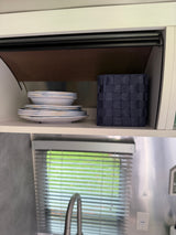 An RV rental's organized kitchen cabinet holds stacked dishes, a storage basket, and reveals a window with blinds and faucet below.