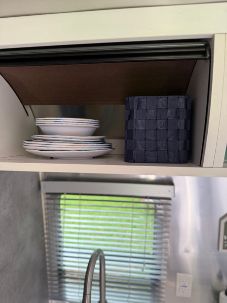 An RV rental's organized kitchen cabinet holds stacked dishes, a storage basket, and reveals a window with blinds and faucet below.