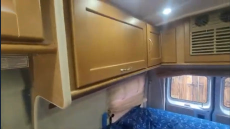 Inside an RV rental: light wood overhead cabinets, bright ceiling lights, a blue patterned bed below, and rear windows showing a fence.